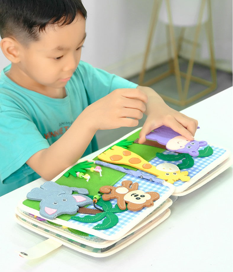 Young boy sitting at table playing with Montessori busy board quiet book, colorful felt activity book with jungle animal pages including elephants and monkeys, sensory learning toy for toddlers aged 12 months and up.